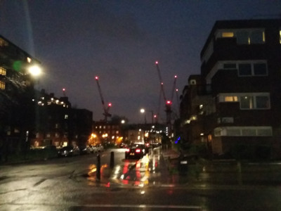 A street of buildings at dusk with several cranes in the distance, red lights glowing at their tips