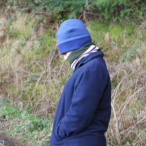 A person standing side-on to the camera in front of a grassy bank. The person is dressed in a blue fleece jacket and hat and has a scarf wrapped around their face so that only a small part of their eyes and nose can be seen.